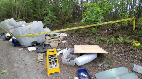 A roadside area in a wooded setting with dumped rubbish, including plastic containers, furniture pieces, and boards, alongside yellow tape and a warning sign.
