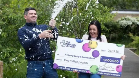 PA Media Canty sprays champagne from a bottle while Sullivan laughs and holds the cheque.