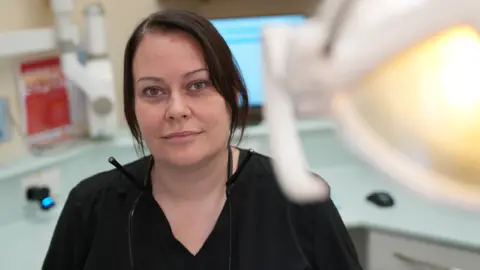 John Fairhall/BBC Hannah Woolnough sits in a dentist appointment room and smiles at the camera. She wears a black dentist uniform and she has dark hair that has been tied back behind her head. 