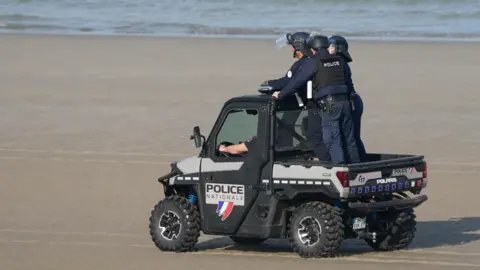 Three French police officers wearing stand on the back of a buggy as it speeds along a beach in Graveline, northern France