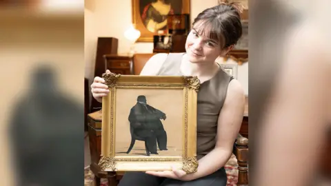 A portrait of a silhouette of the man once known as England's fattest is held by Holly Hammond from Duggleby Stephenson auction house. The painting is in a gold frame. 