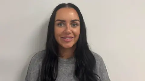 BBC A girl with dark brown hair stands against a white wall. She is smiling, teeth showing as she wears a grey woollen jumper.