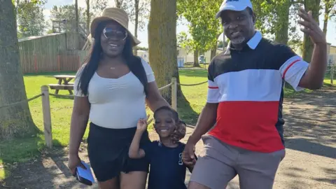 supplied A man and woman and young son hold hands and smile in the sunshine. They are all dressed in summer clothes