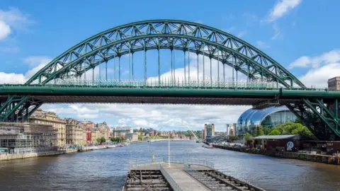 Gateshead Council Restored section of the Tyne Bridge showing scaffolding and the structure's arch, freshly painted in the shade Hollybush Green. The Tyne River is below and it is a clear, sunny day. 