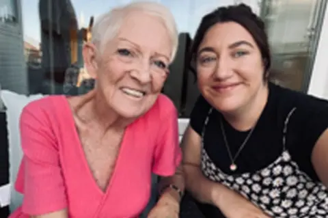 Ellie Atkins-Tate Two women smiling at the camera. One on the left has a pink top and short white hair, the other has dark hair and is wearing a black t-shirt under a flowery dress.