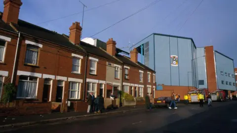 Getty Images Highfield Road stadium before demolition stood next to homes. The stadium is next to a row of terraced houses and has a blue wall with the club's crest on it.