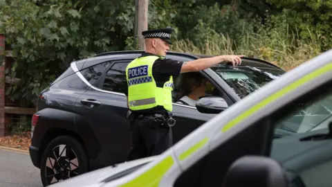 EPA A male police officer in a black and white hat, black top and trousers and a yellow hi-vis vest that says "police" on the back. He is standing next to a black car and is talking to the driver, pointing off into the distance to direct them