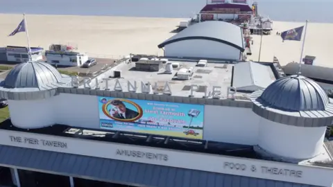 Jamie Niblock/BBC A top view of the Britannia Pier with its cream façade and grey turrets. The theatre down towards the bottom of the pier has burgundy signage. The sandy beach is on either side of the pier and the sea is in the background. 
