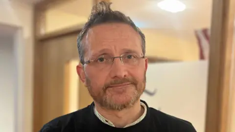 Donal is smiling at the camera. He has grey hair and is wearing a white shirt, black jumper and glasses. 