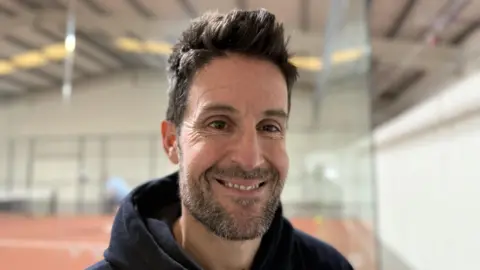 Ryan smiles for the camera at his padel club. A court is seen behind and he is wearing a black hoodie and has stubble on his chin.