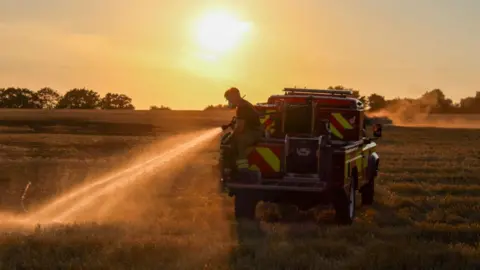 HIWFRS/Ashley Simmonds Firefighter with water spraying from a hose sat on a 4/x fire appliance in a field which is smoking. The sun is setting.