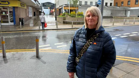 BBC/Elliott Green A woman with blond hair in a bob is wearing a navy puffer jacket with a crossbody bag and is standing in a street