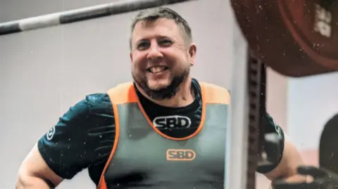 Chris Pryor A head and shoulders picture of Chris Pryor wearing a black t-shirt and orange and grey athletic vest. He is standing with his hands on his hips and smiling. There is a large barbell out of focus in the foreground.