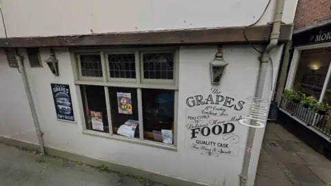 The outside of the Grapes Tavern. It is a white building with the name printed on the side in black lettering. The window contains posters for events, and there is an advert for burgers to the left of the window.