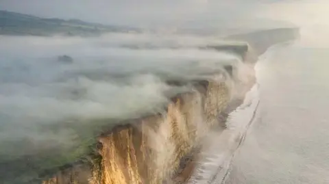 Thick fog rolls off the top of cliffs and into the sea. The sky is grey and foggy. The cliffs have a beige colour to them. A calm sea is rolling onto the shore at the foot of the cliffs. 