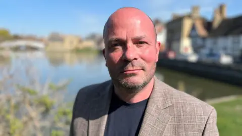 Phillip Malley looking into camera in front of the waterfront in Godmanchester. He has stubble and is wearing a brown suit and black t-shirt.