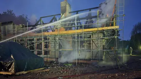 Smoke comes from timber frame of roof of house partially built - it is surrounded by scaffolding and is built with large grey blocks/bricks.
