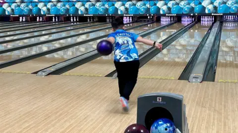 Stroud Sharks YBC Mylo wearing the branded blue and white bowling shirt with a shark on it at the Stroud Bowl. He is bowling a purple ball down the lane towards 10 pins.