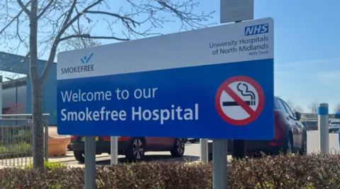 A mainly blue and white sign by a hedge includes the words "Welcome to our Smokefree Hospital" and a red, white and black no-smoking sign. Some cars are visible in the background.