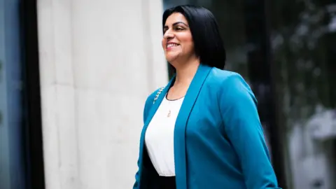 Shabana Mahmood smiles as she walks past a beige brick building wearing a blue suit and white shirt as she arrives at the BBC building in London on Sunday.