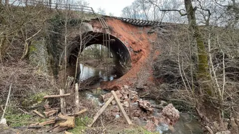 BBC A bridge underneath a railway with a river running underneath it. Earth has fallen away from the railway track leaving it exposed. There is debris and fencing in the river below.