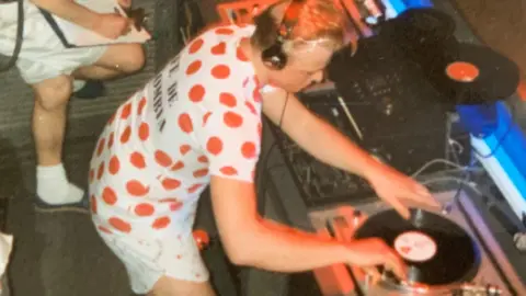 Paul Stainton Paul Stainton cues up a record. He is wearing a red polka-dot cycling shirt and shorts. He has blonde hair and is wearing headphones.