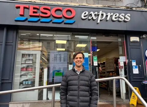 A man in a black puffer coat stands in front of a Tesco store, smiling.