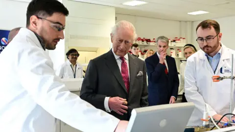 King Charles stands in between two men in white lab coats while all three look at the screen. The King is wearing a grey suit with a white collared shirt and a red patterned tie. There are other scientists in the background.