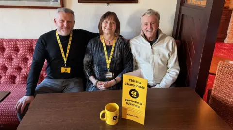 Jon Haslam A man in a black jumper with a black lanyard sits next to a woman with a yellow lanyard and a man in a cream jacket. They are sat at a dark wood table smiling at the camera. A yellow mug and leaflet is on the table. 