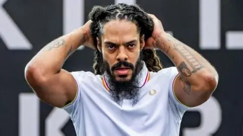 A man performing on stage during a Bob Vylan set. He is wearing a white polo shirt and his hands are touching his head. He has a dark beard and dreadlocks and tattoos on both his arms.