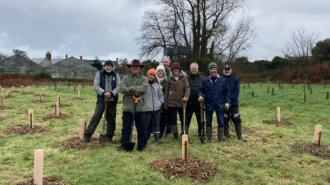 BBC News A white man in a green coat and a brown cowboy hat, stands in front of a group of people in coats, leaning on a shovel. 