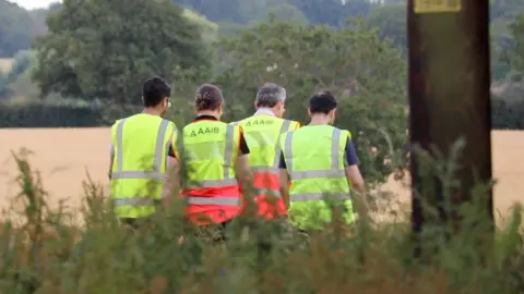 Stuart Woodward/BBC Four investigators wearing hi-vis vests while looking at something in a field. They walk away from the camera. In the foreground is green foliage.