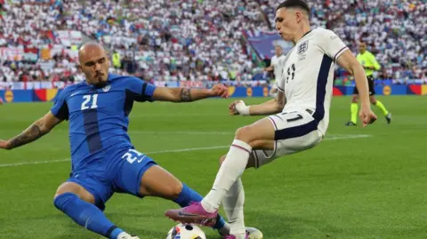 Getty Images England footballer Phil Foden on the left wing in an all white playing kit being tackled by Slovenia's defenderby a 