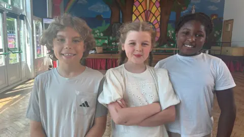 (L-R) Emanuel, 10, Amelie, 10, Olanna, 9. They all smile at the camera stood inside a school hall.