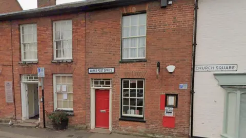 A red brick building with a sign saying Bures Post Office. It has a red door and two windows. There is a post box on the outside of the building. There are other buildings either side of the post office.