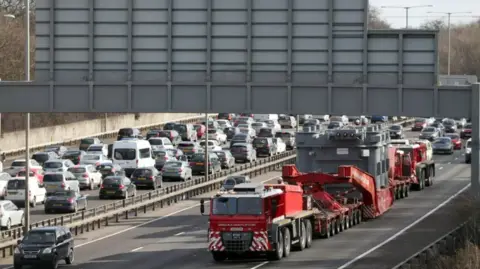 PA Media A long, wide, red lorry carrying a huge transformer travels down a very busy motorway 