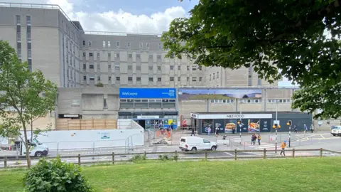 The exterior of Derriford Hospital. There is a large blue sign above the entrance doors that reads WELCOME with NHS branding. There is a single storey M&S Food shop to the left of the entrance. A road runs parallel to the building with a pedestrian crossing on it. In the foreground is a fenced off area of grass with a couple of small trees.
