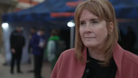 Policing Minister Sarah Jones wears a pink woollen jacket, black top. She has light brown hair, a fringe and looks serious. The background is blurred but people are stood about chatting.