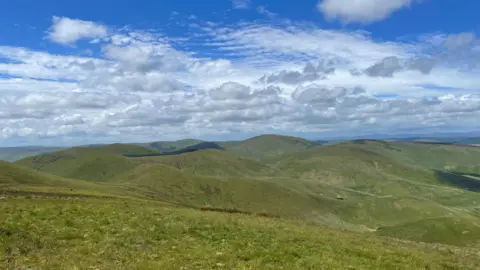 Muirhall Energy A set of rolling hills with low clouds and a blue sky above it where a wind farm could be built