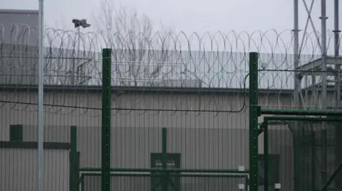 High fences with circled barbed wire at the top. A large metal building is on the other side.