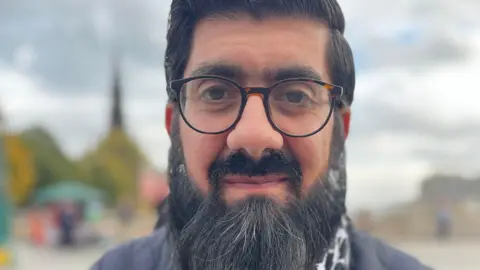 Omar Afzal, A Muslim man with black hair and a beard. He is wearing black glasses.