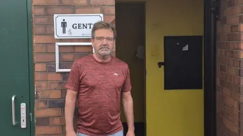 Paul White stands beside the entrance to the public toilet. It is a red brick building with yellow interior walls. There is a sign above White's head that reads: "Gents" and a green door on his left. He is wearing a dark red shirt and looking solemnly at the camera.