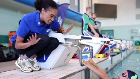 Alice Dearing crouches down by a starting block at a swimming pool, talking to a young swimmer in the water