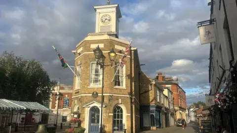The photo shows a historic building which is made of yellow/brown brick. It has Georgian-style windows and a clock face at the top. It's set in a high street.