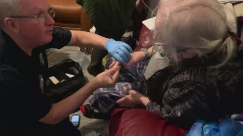The picture shows a paramedic providing medical care to an elderly person in their own home. The paramedic is wearing protective gloves and has a medical kit open beside them on the floor, along with monitoring equipment. The setting looks like a living room, with everyday household furniture around them, suggesting the care is being delivered in the community rather than in hospital.