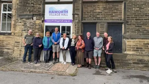 A mixed gender group of people stand outside a tall, brown brick building with a white sign affixed that reads in purple lettering Haworth Village Hall. 