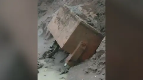 A red brick rectangular structure lays against a cliff face on a beach.