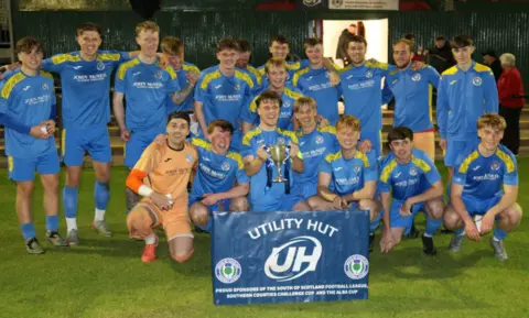David McDonald A group of footballers in blue strips with yellow shoulders hold up a cup and celebrate their win