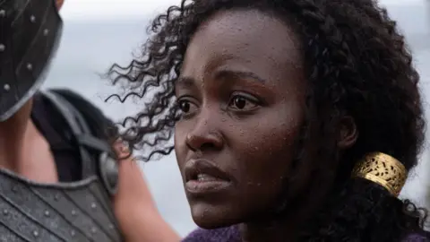 Lupita Nyong'o looks to the left of frame in a concerned manner, in a close head and shoulders shot as she stars as Helen of Troy in Christopher Nolan's The Odyssey.