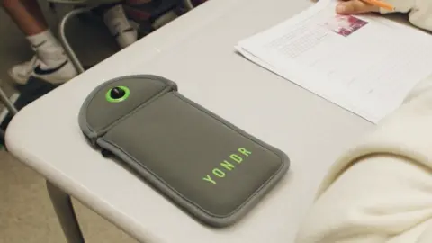 Grey phone pouch with YONDR written on it in green lettering laying on a pupil's desk